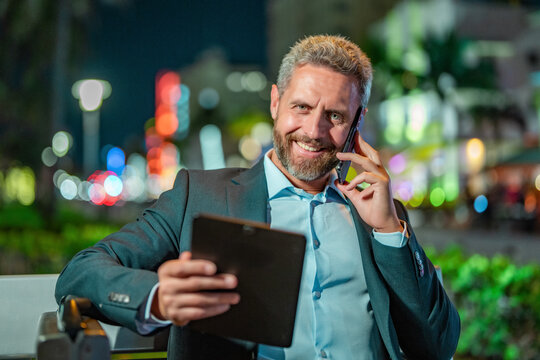 Man in business suit talking on phone and using tablet. Business man with tablet outdoor. Business man with a tablet and phone outdoors. Businessman using tablet, sit on bench. Night business.