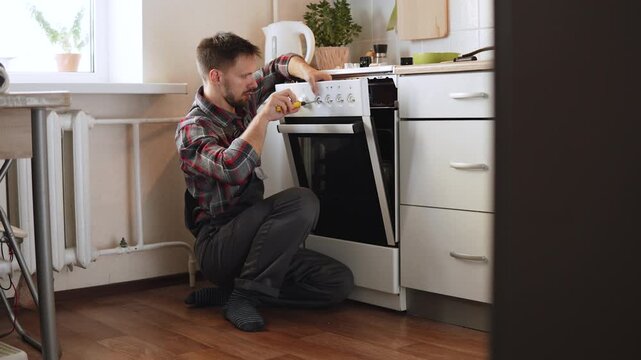 Appliance repair technician fixes electric stove in apartment kitchen, push out shot. Handyman disassembles panel to troubleshoot problem, gimbal wide shot.