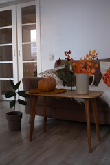 This cozy living room with a pumpkin on wooden table, plants in pot, sec autumn branches in ceramic vase and modern furnishings creates an inviting autumn atmosphere.