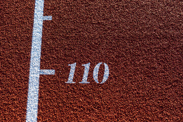 Close up photo of outdoor running track lane white text noting the 110, distance, part of a long jump, triple jump, on a new red track with white lane lines.