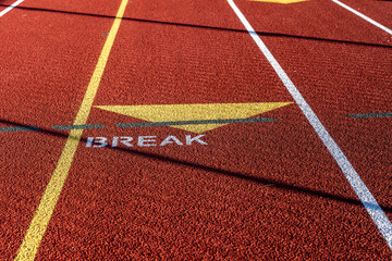 Close up photo of outdoor running track with white and gold lane line, with the word break, with exchange zone triangle, on a new red track.