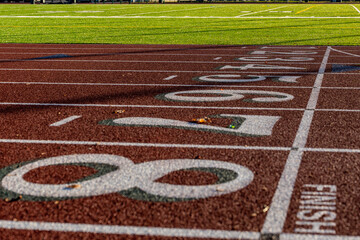 Close up photo of outdoor running track lanes with white numbers one, two, three, four five, six, seven, eight, 1, 2, 3, 4, 5, 6, 7, 8, with green shadow on a new red track with white lane lines.
