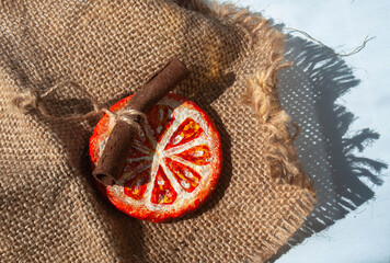 Hand-made Christmas papier mache ornament painted with acrylic paints. Round dried orange slice with cinnamon stick on a jute thread lying on Hessian fabric piece. DIY home decoration project. Craft h