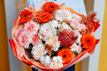Warm autumn bouquet of orange gerbera daisies, blush pink roses, white cotton pods, and red leucospermum (pincushion protea), held by a man