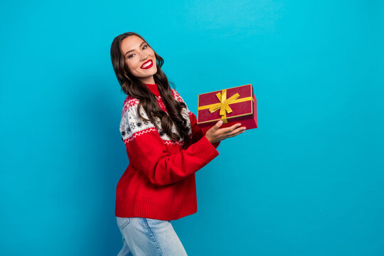 Cheerful woman in festive sweater holding gift box against blue wall, celebrating Christmas season with excitement and joy