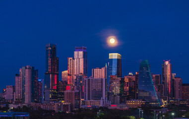 Naklejka premium Aerial drone view of the Austin Texas skyline as the full harvest moon is seen rising by the office buildings. Zilker Park in the foreground.