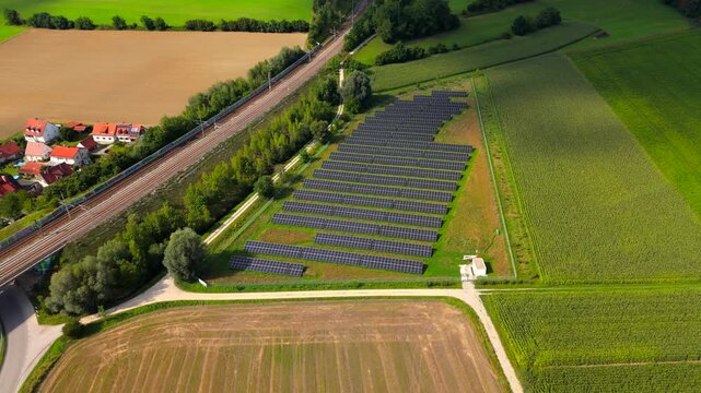 Luftaufnahme mit Drohne uber Bayern, Deutschland. Bahngleise fuehren durch Felder vorbei an einer Solaranlage. Solarpaneele glaenzen in Sonne und zeigen nachhaltigen Verkehr und erneuerbare Energie