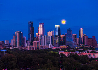 Naklejka premium Aerial drone view of the Austin Texas skyline as the full harvest moon is seen rising by the office buildings. Zilker Park in the foreground.