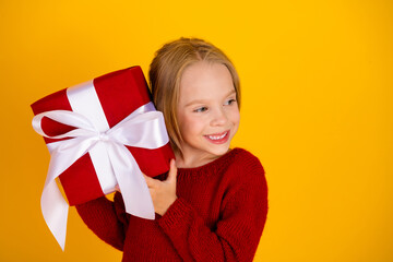 Festive kid girl with large red gift and white ribbon smiles in cozy red sweater against bright yellow background for a joyful Christmas moments