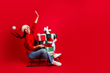 Festive young woman celebrating the holiday season on a sleigh with gifts against a vibrant red...