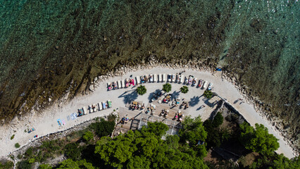 Aerial view of a rocky beach with clear turquoise water, sunbeds, umbrellas, and people relaxing near the shore surrounded by green trees on a sunny day