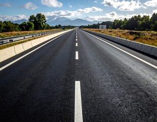 Fototapeta premium A vast highway stretches ahead, disappearing into the distant mountains under a bright blue sky with fluffy clouds. Lush greenery lines the sides