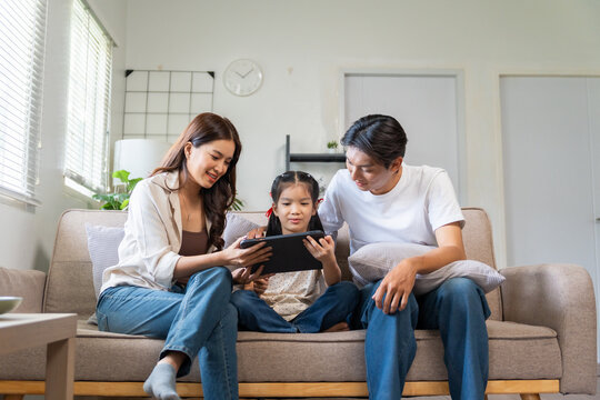 Happy Asian family spending bonding time. Parents and daughter sitting on couch, watching and learning on digital tablet