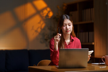 Focused young Asian woman working on laptop and taking notes, studying or remote working from home during the night