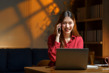 Smiling young adult asian woman working remotely at home office, using laptop and talking on mobile phone