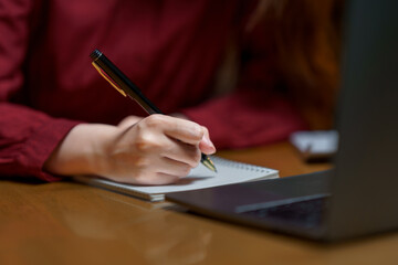Woman writing notes with pen on notebook while studying at a desk with a laptop, focusing on learning and education