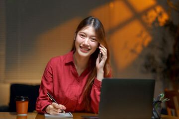 Young woman working late, smiling while managing a phone call and writing notes at desk with laptop and coffee