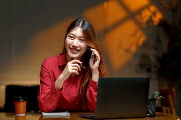 Asian businesswoman smiling, holding a pen, and talking on her mobile phone while working on a laptop at her desk