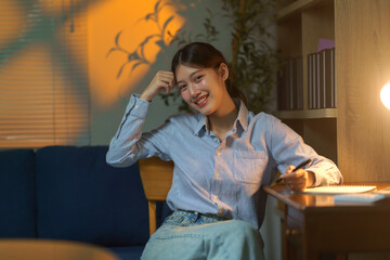 Young Asian woman student smiling, studying in a home library at night, sitting at a desk with a lamp