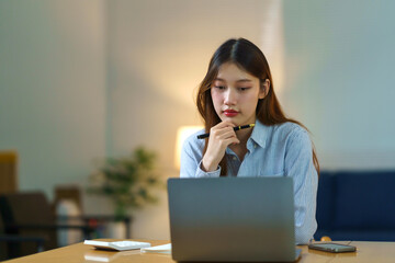 Focused young Asian woman concentrating, holding pen, working on laptop at home office desk, representing remote work and studying