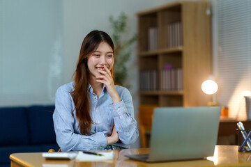 Young woman laughing, covering mouth during a video call on a laptop while working from her home office desk