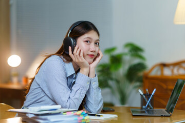 Young Asian woman wearing headphones, sitting at desk with laptop, studying or working remotely from home, looking at camera