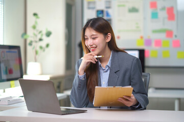 Asian businesswoman smiling, holding a pen and clipboard, participating in an online video call from her modern office desk