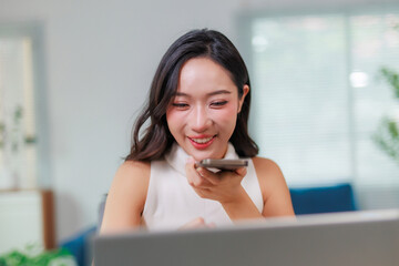 Young Asian woman speaking into her smartphone, utilizing voice assistant technology while working on a laptop in a modern office
