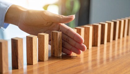 "Row of upright wooden dominoes on polished surface, hand intervening mid-sequence to stop falling motion, symbolizing prevention, control, and breaking a chain reaction."