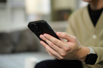Close up of a man holding a smartphone and typing on the screen while sitting on a couch in a living room, for business, communication and technology concept.