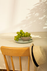 Cozy summer still life with green grapes, wooden chair and straw hat in sunlight and shadows