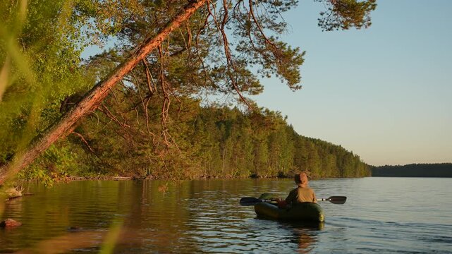 A man kayaks across a quiet forest lake at sunset. A hat-wearing traveler paddles a canoe along the lakeshore beneath an overhanging tree. A man on a packraft drifts across the lake, taking in the