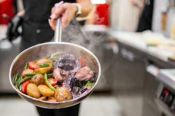 Chef holding hot frying pan with steaming seafood and vegetables in professional kitchen. Colorful dish includes octopus, potatoes, beans, and tomatoes. Culinary preparation concept.