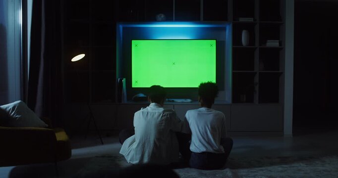Man and woman playing video game while sitting in front of green screen TV