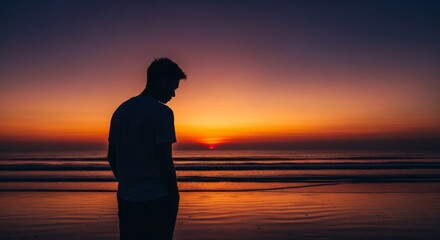 Fototapeta premium Silhouette At Sunset On Beach - A person stands silhouetted on a beach, watching the sunset over the ocean. The sky is filled with warm colors