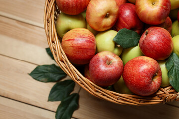 Fresh apples in wicker basket and green leaves on wooden table, top view. Space for text
