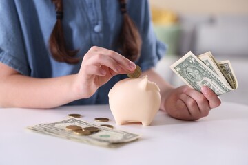 Little girl putting coin into piggy bank at white table indoors, closeup. Pocket money and responsibility