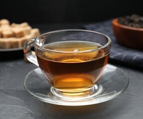 Aromatic tea in glass cup, brown sugar and dried leaves on black table, closeup