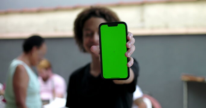 Young boy holding smartphone with green screen chroma key, smiling outdoors in casual courtyard setting, people blurred in background