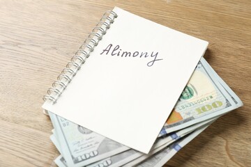 Notebook with word alimony and money on wooden table, closeup