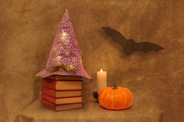Halloween still life with several red books, a witch hat, a pumpkin, a white candle, and a bat, on a brown background