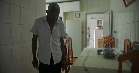 One Elderly Black man walking from kitchen to living room, wearing a neutral expression,...