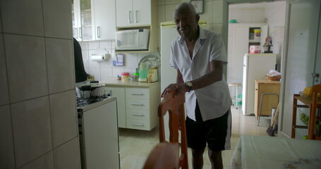 One Elderly Black man standing in kitchen near dining chair, appearing thoughtful, with a homely...