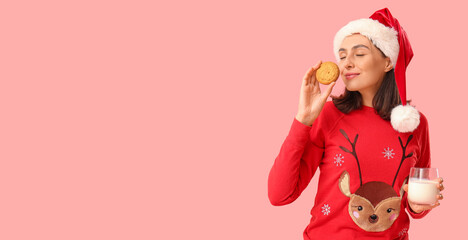 Beautiful happy young woman in Santa hat with cookie and glass of milk on pink background