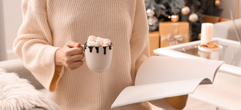 Woman holding cup of hot chocolate with marshmallows while reading magazine in festive living room