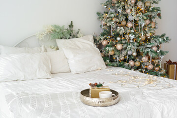 Elegant gift box and ceramic cup on silver tray with Christmas tree lights on cozy white bed in festive decorated bedroom interior. New Year and Christmas atmosphere, festive home decor for holidays