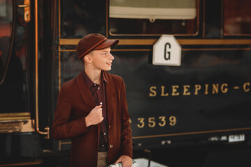 A thoughtful young man in a brown coat and a retro-style cap holds a rolled-up newspaper on a train...