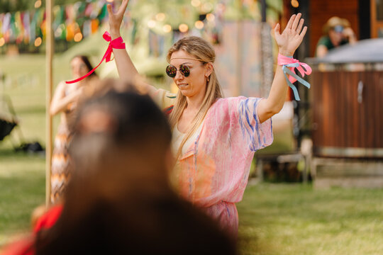 Woman in colorful boho outfit and sunglasses raises her hands while dancing at an outdoor hippie festival, with vibrant ribbons and decorations in the background.