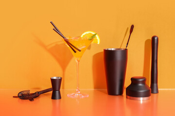 Bartender utensils and glass of cocktail with citrus slice on orange background