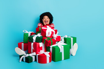 Excited young woman sitting among multiple colorful gift boxes against a vibrant blue background, radiating holiday joy